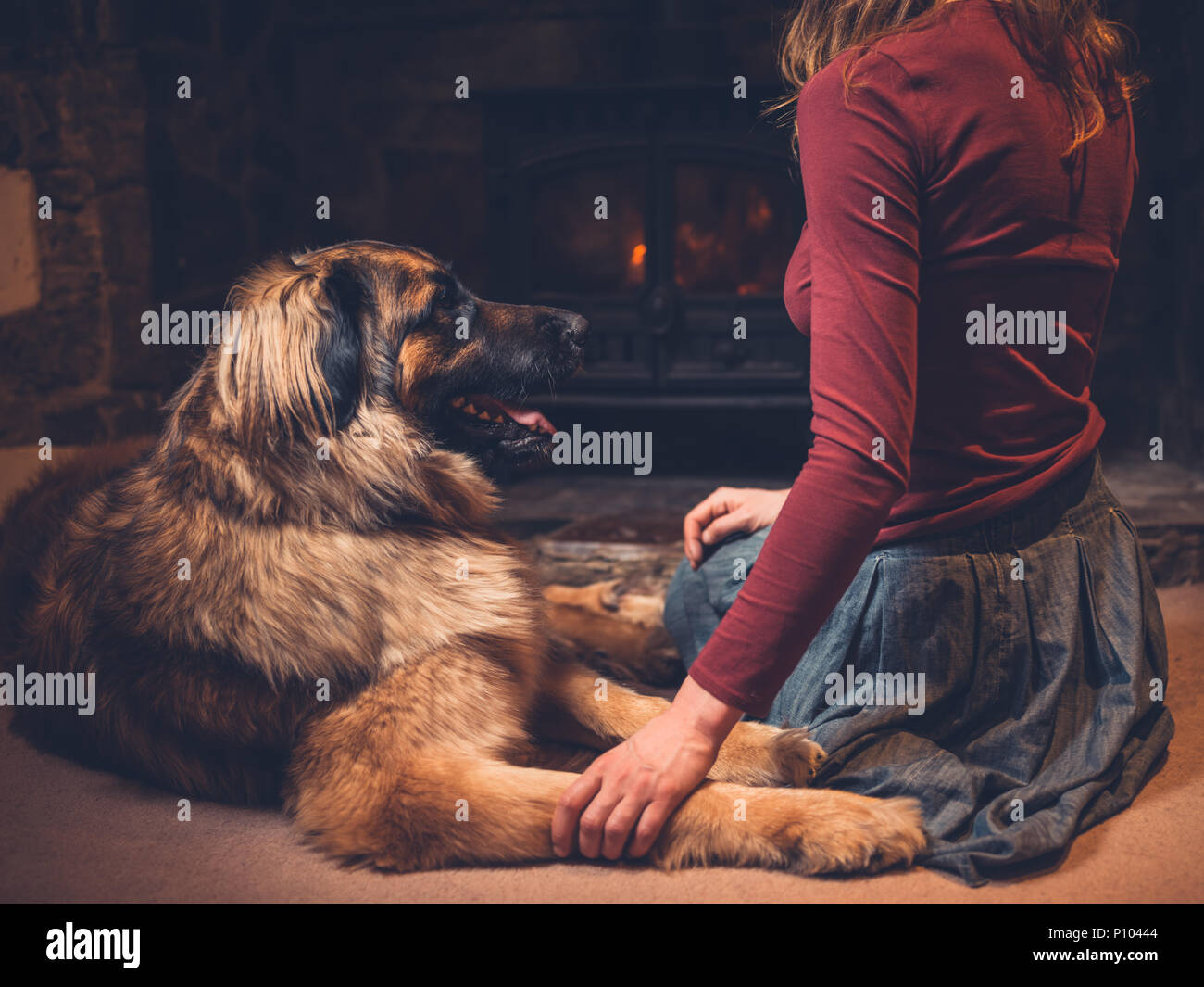 A young woman is sitting by the fire of a log burner with a giant dog