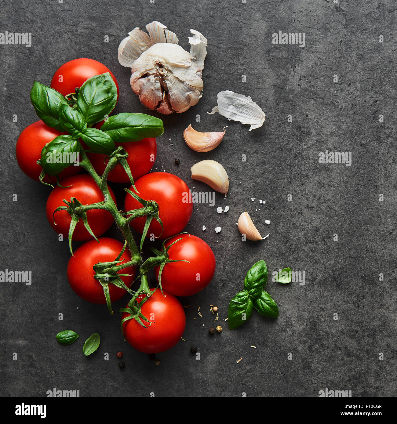 Food background. Flat lay of fresh tomatoes with basil, garlic and sea salt on black stone background. Square crop, top view. Stock Photo