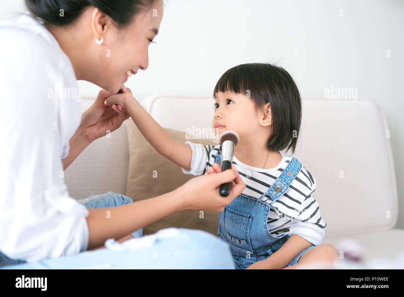 Cute little girl and her beautiful mother are doing makeup while sitting on couch at home Stock Photo