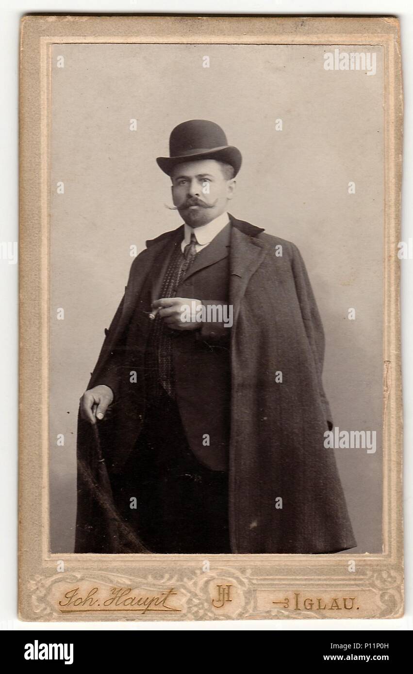 JIHLAVA, AUSTRIA-HUNGARY - CIRCA 1905: Vintage cabinet card shows dandy man wears bowler hat, luxury garment and holds walking stick and cigar. Antique black white photo. Stock Photo