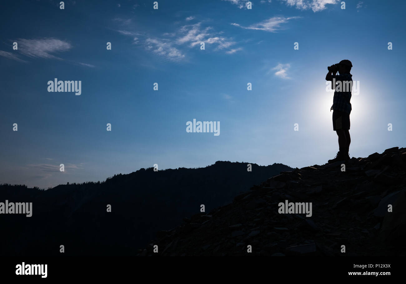Hiker wearing shorts stands on mountain top with binoculars or camera, backlit by the sun. Stock Photo
