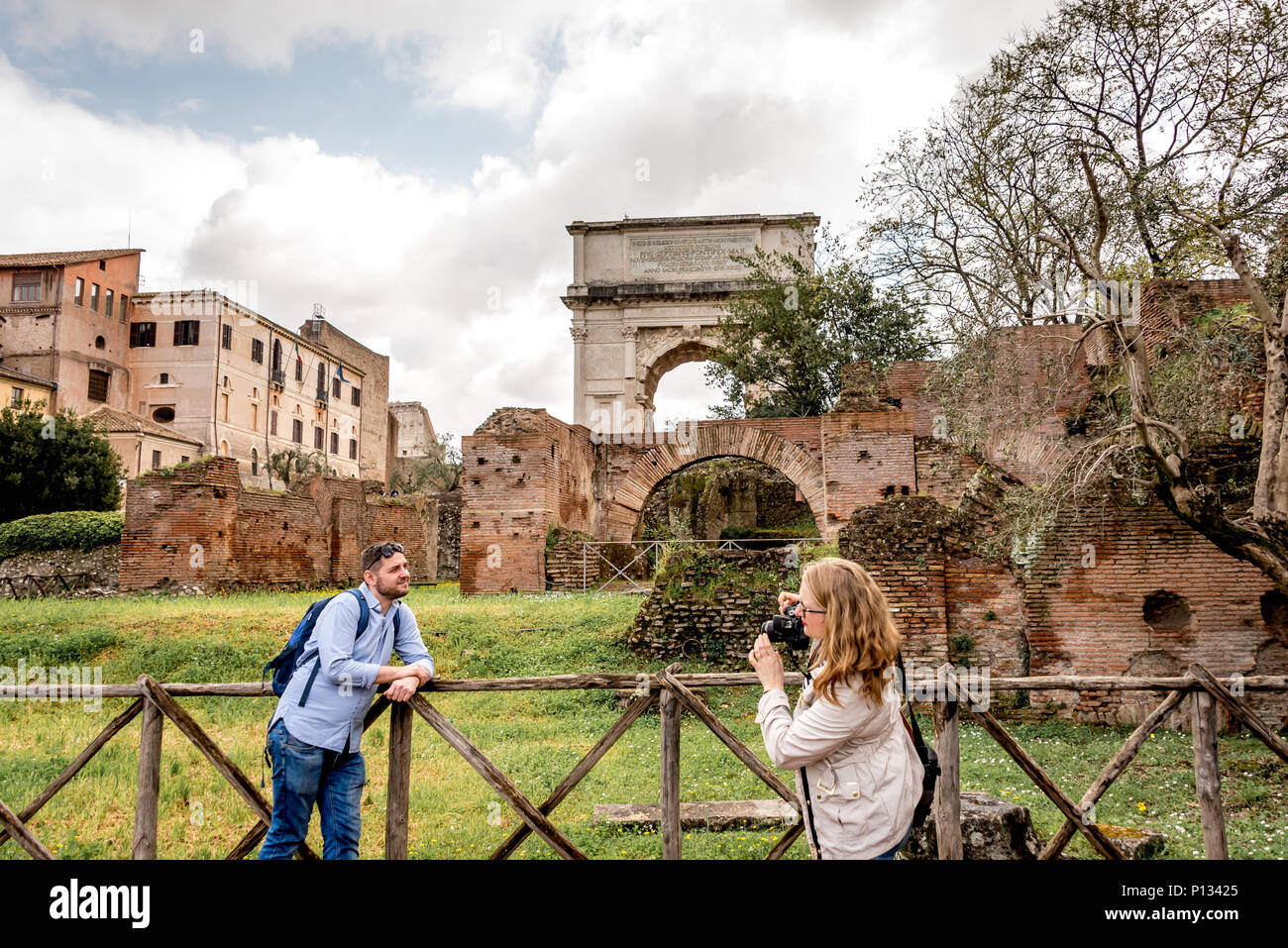 Roman Forum, a man poses for a photo among the ancient ruins found in the city center, a woman holds the camera as he leans on fence for a picture. Stock Photo