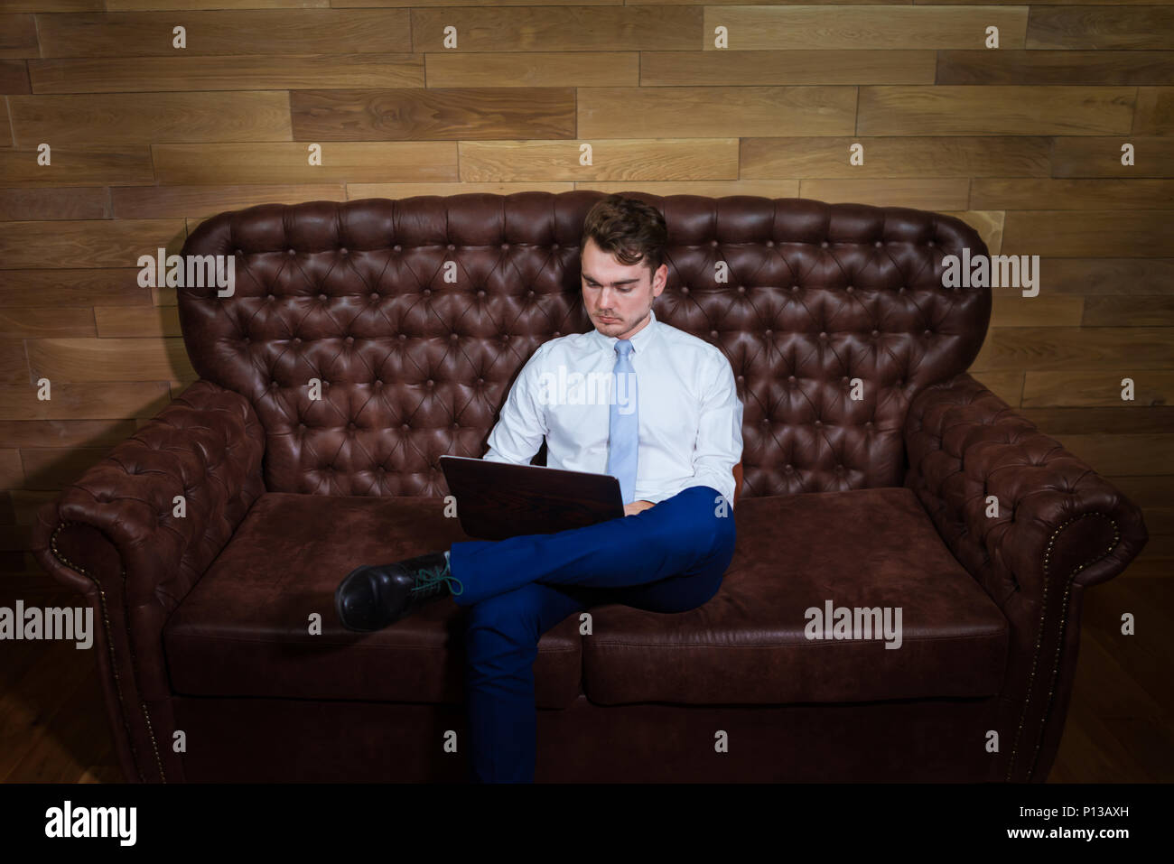 Serious man in suit is working with laptop at home office. Confident young manager sits on the leather sofa on background of wooden wall. Stock Photo