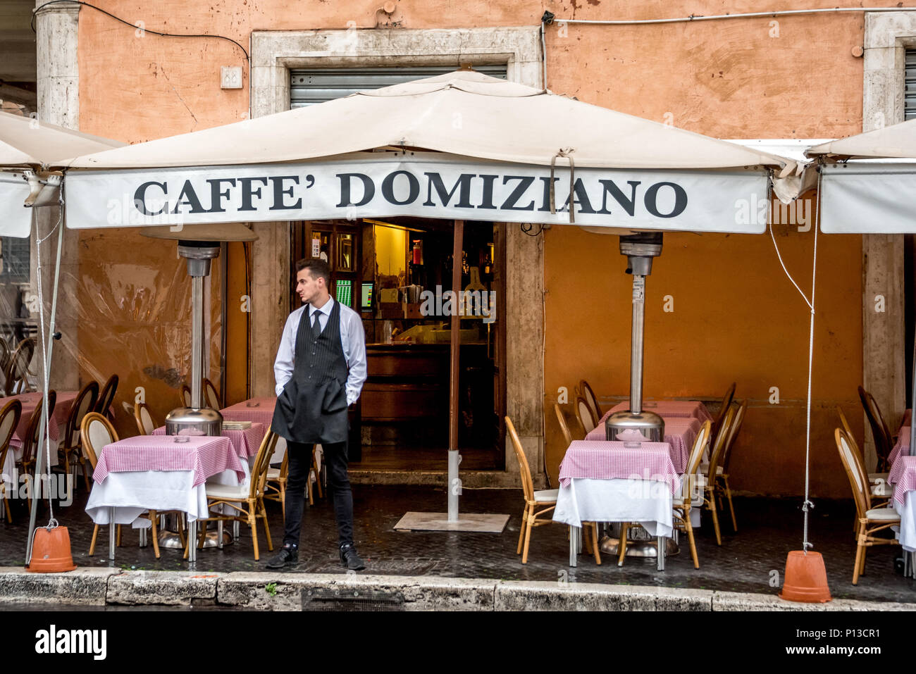Italian waiter stands outside Caffe Domiziano restaurant on Piazza Navona waiting for customers on a rainy day with empty tables for outdoor dining. Stock Photo