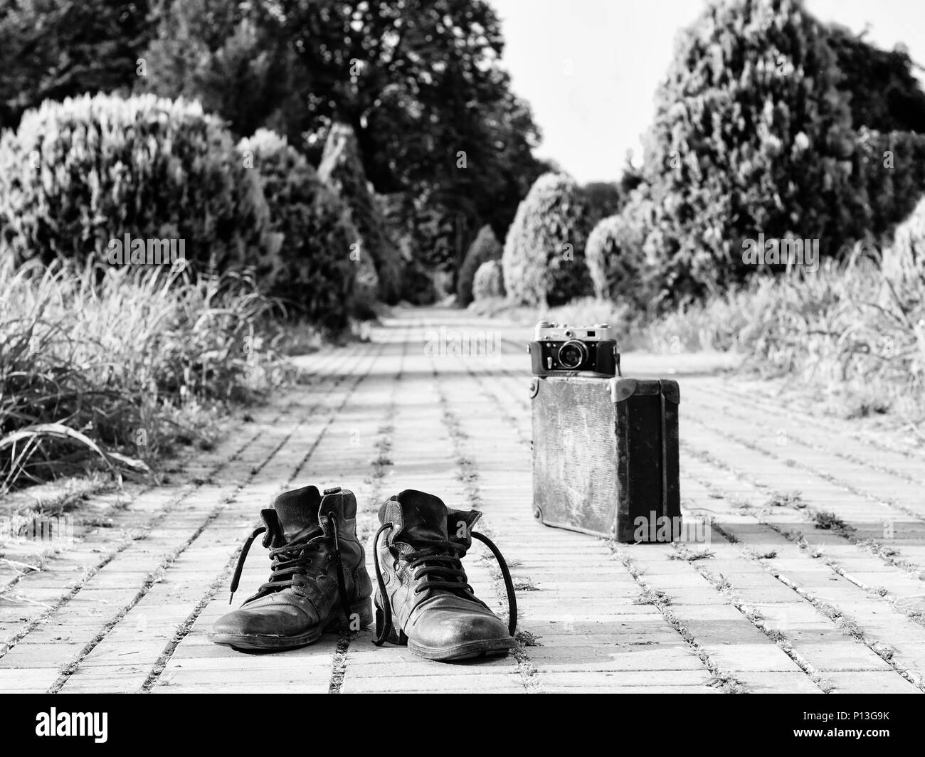 Leather boots in a foreground, in the middle of a brick road. A film camera on an antique suitcase, in a blurry background. Black-and-white photo. Stock Photo