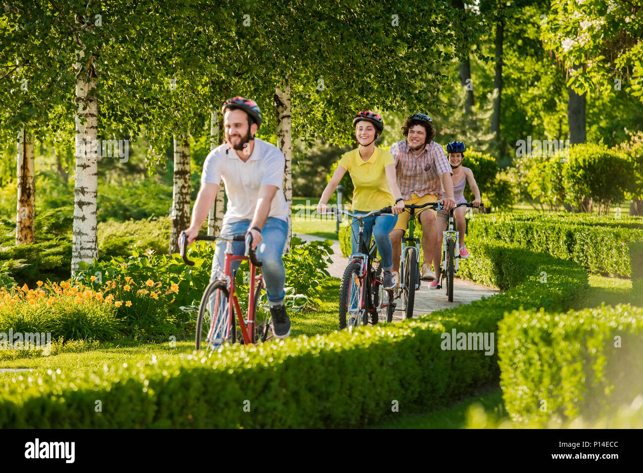Happy friends cycling in city park. Group of students riding bikes on summer nature background. People and active rest. Stock Photo