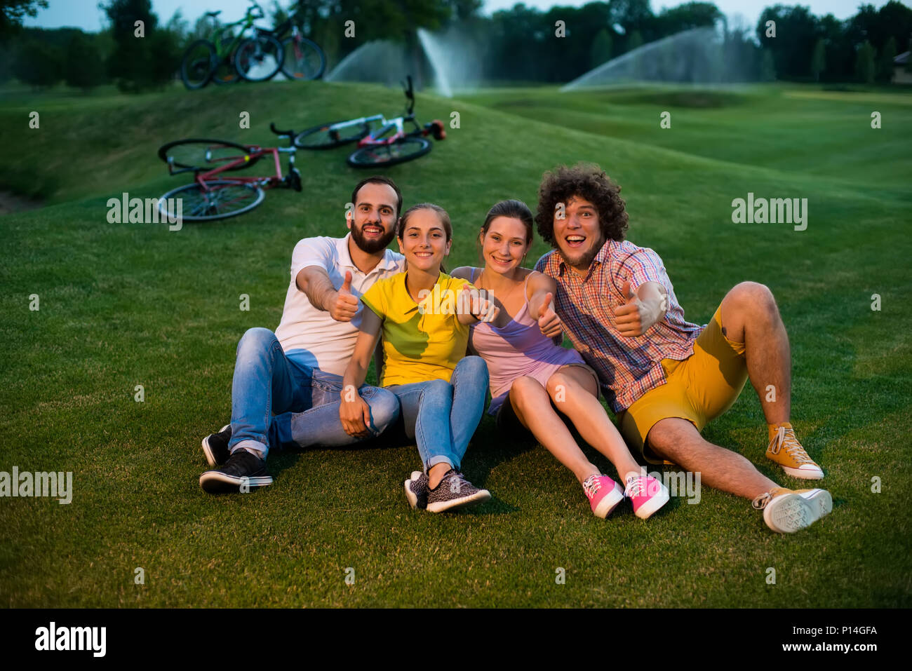 Group of friends gesturing thumbs up outdoors. Four cheerful cyclists sitting on the grass and showing thumb up gesture. Best weekend with friends. Ev Stock Photo