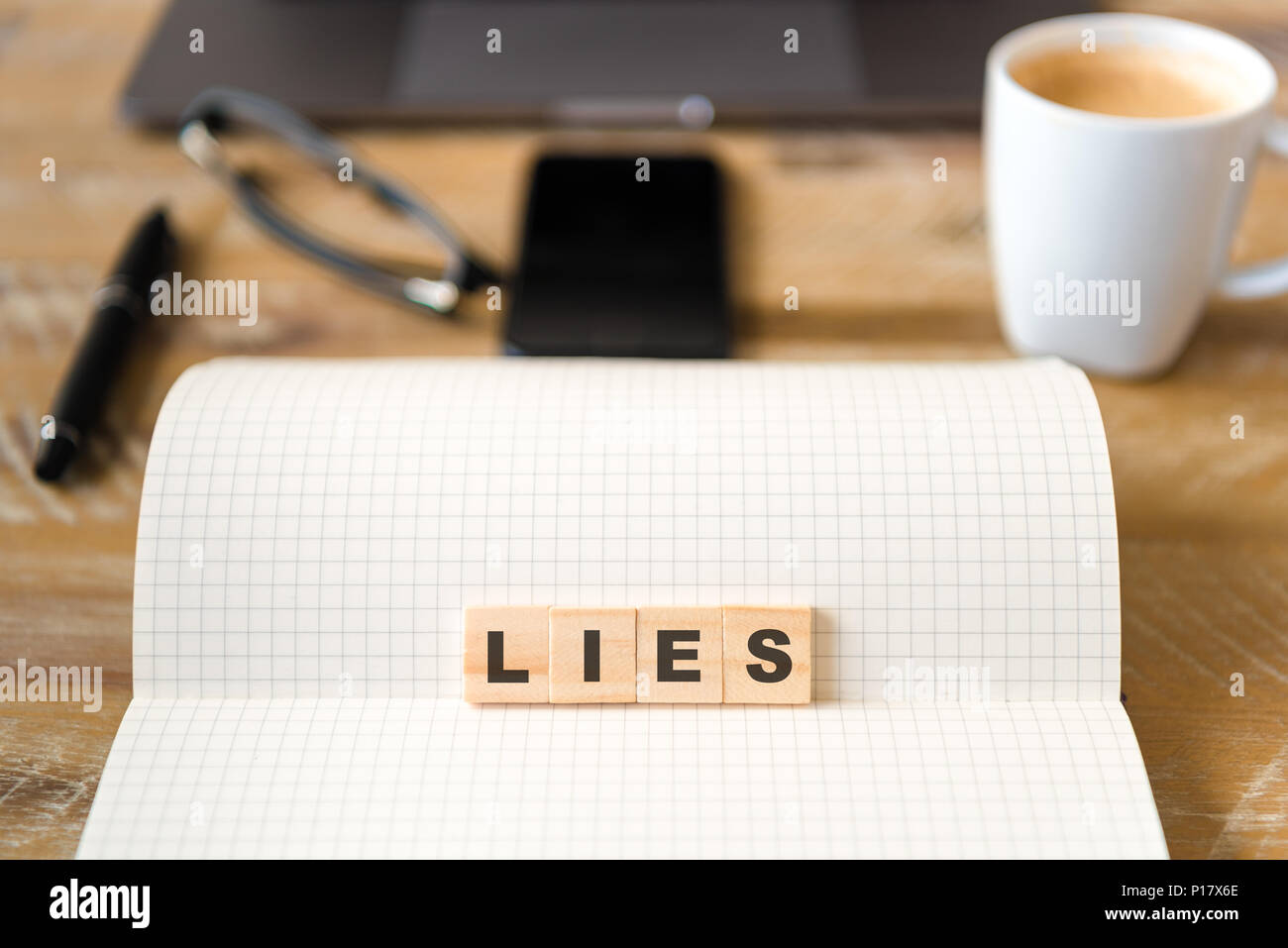 Closeup on notebook over vintage desk surface, front focus on wooden blocks with letters making Lies text. Stock Photo