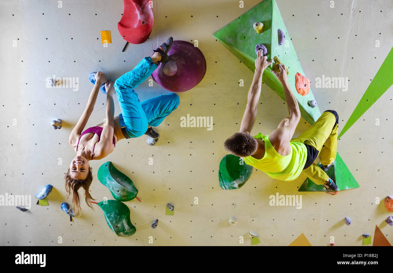 Rock climbers in bouldering gym climbing up overhanging wall. Young woman hanging upside down and smiling, male climber looking at her. Stock Photo