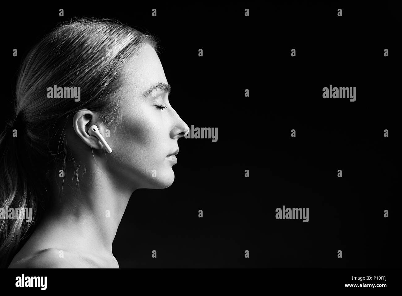 profile of blonde young woman with bluetooth earphones and closed eyes on black background, monochrome Stock Photo