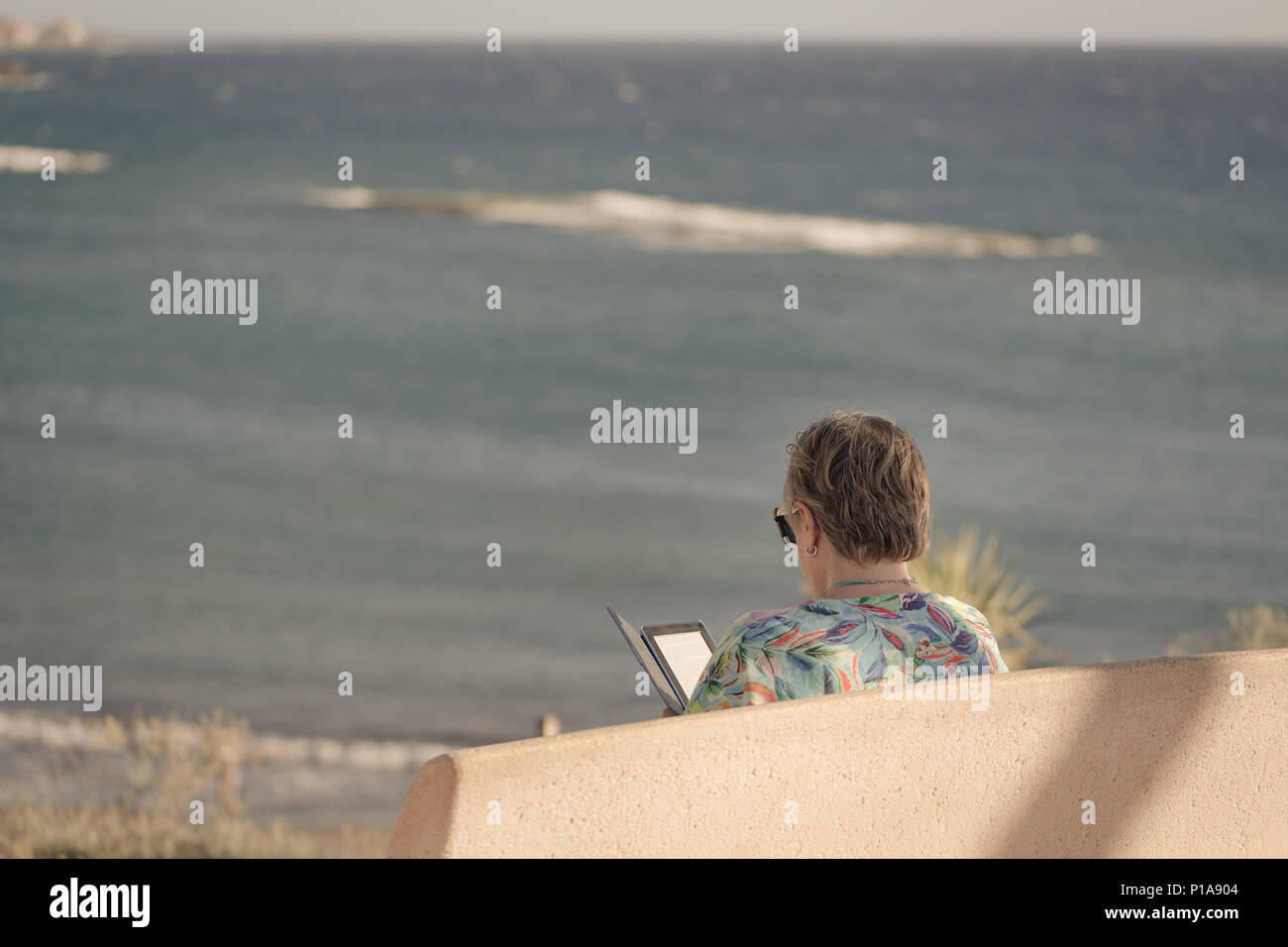 Older woman is sitting on bench with view on sea and she is reading ebook on her reader. Stock Photo