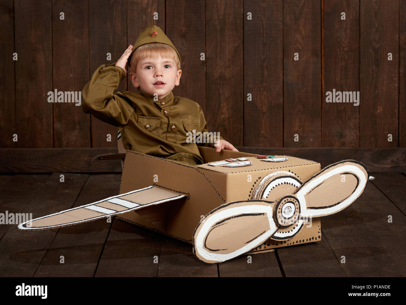 children boy are dressed as soldier in retro military uniforms sit in an airplane made of cardboard box and dreams of becoming a pilot, dark wood back Stock Photo