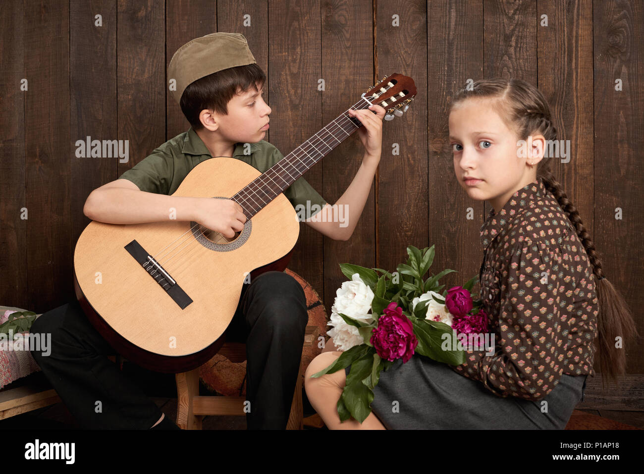 children are dressed in retro military uniforms sitting and playing guitar, sending a soldier to the army, dark wood background, retro style Stock Photo