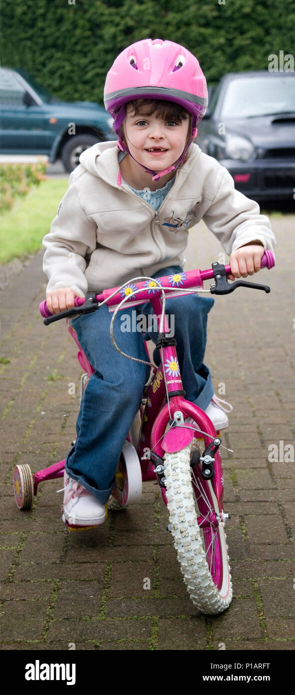 Little girl with front teeth missing happily playing on her pink bicycle with stabilisers and pink helmet looking towards camera and smiling. Stock Photo