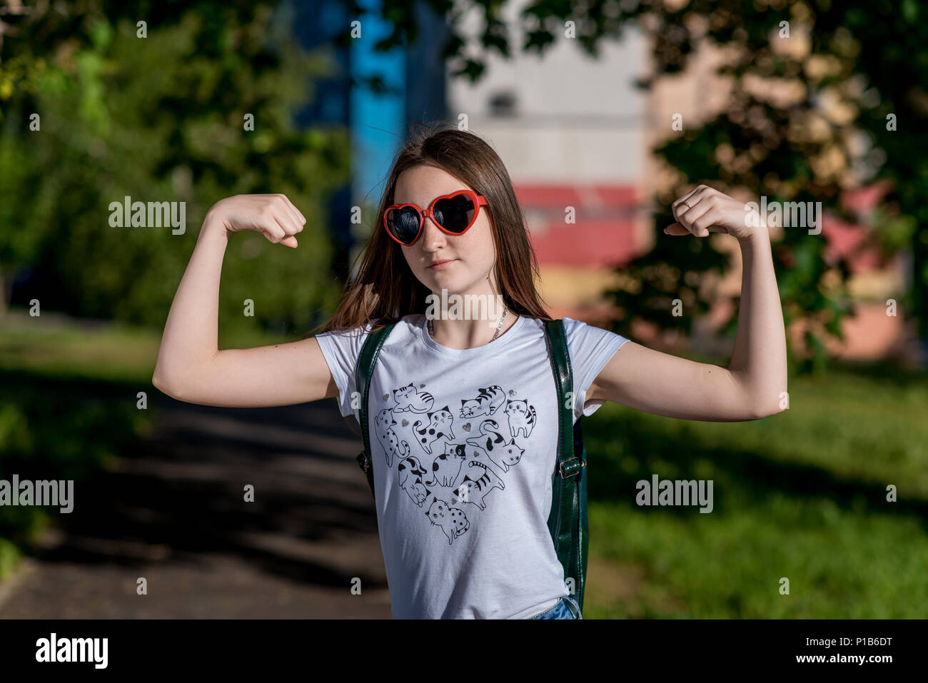 A teenage girl jokingly shows her biceps on her hands. A bright sunny day in the park. In sunglasses in form of red hearts. Emotions of the power of female courage. Teenager after school. Stock Photo