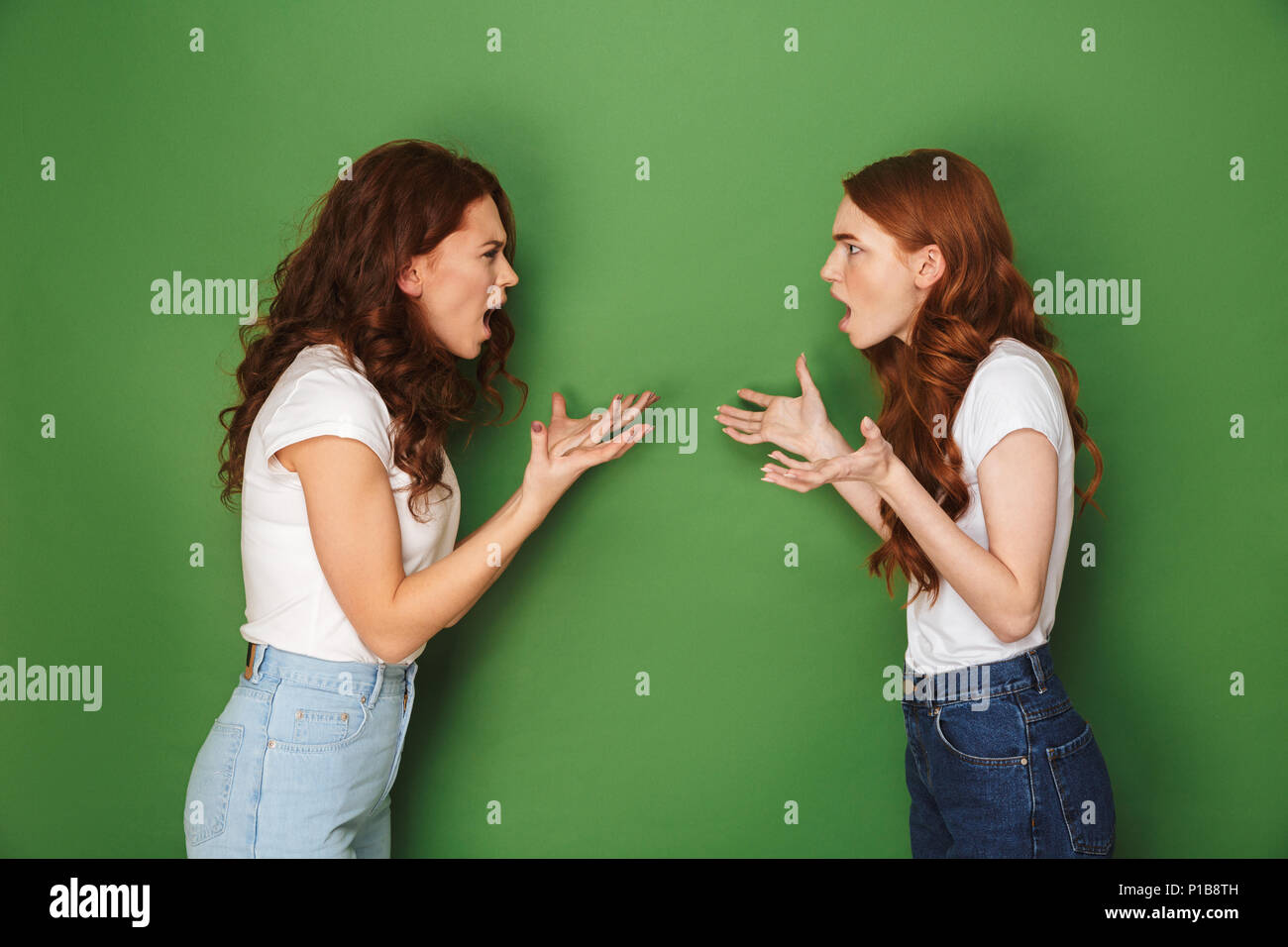 Two outraged girls 20s with ginger hair standing face to face crossed and shouting at each other isolated over green background Stock Photo