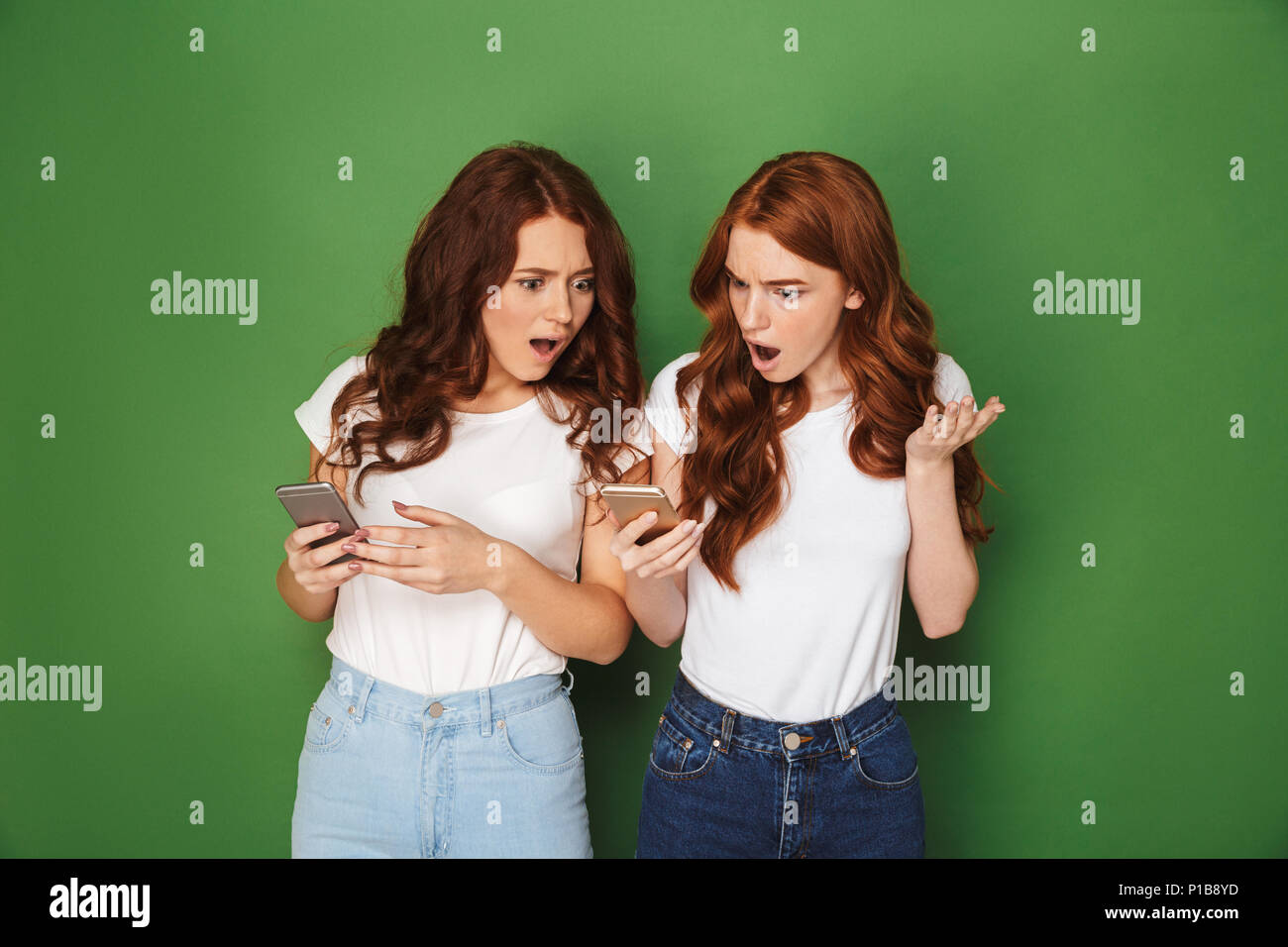 Portrait of two outraged women with red hair using cell phones and expressing indignation isolated over green background Stock Photo
