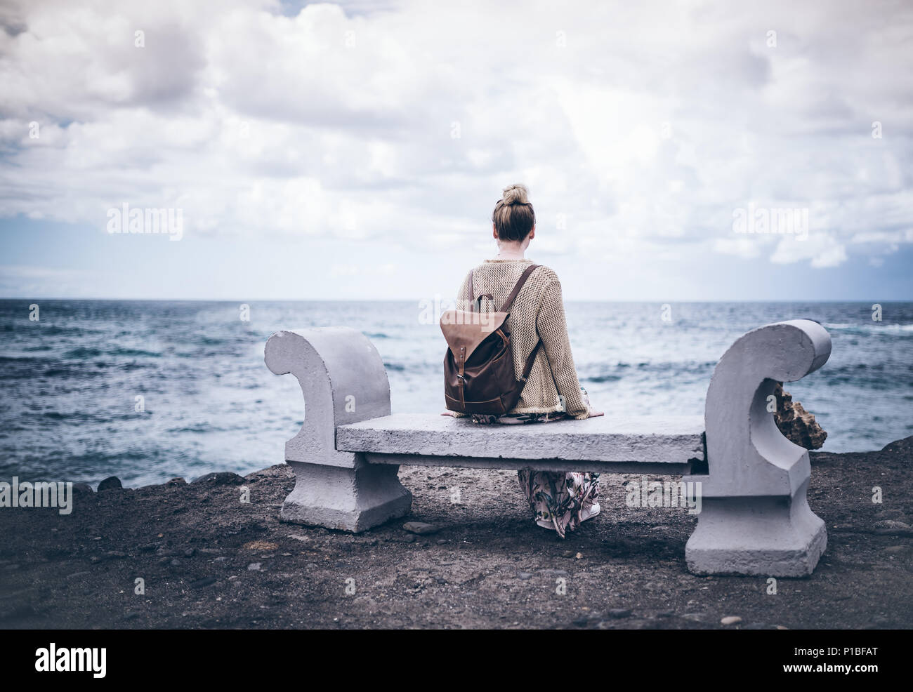 back view of young woman in long dress and with backpack sitting on stone bench by sea Stock Photo