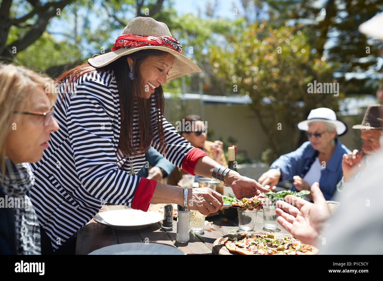 Smiling senior woman reaching for pizza at sunny garden party table Stock Photo