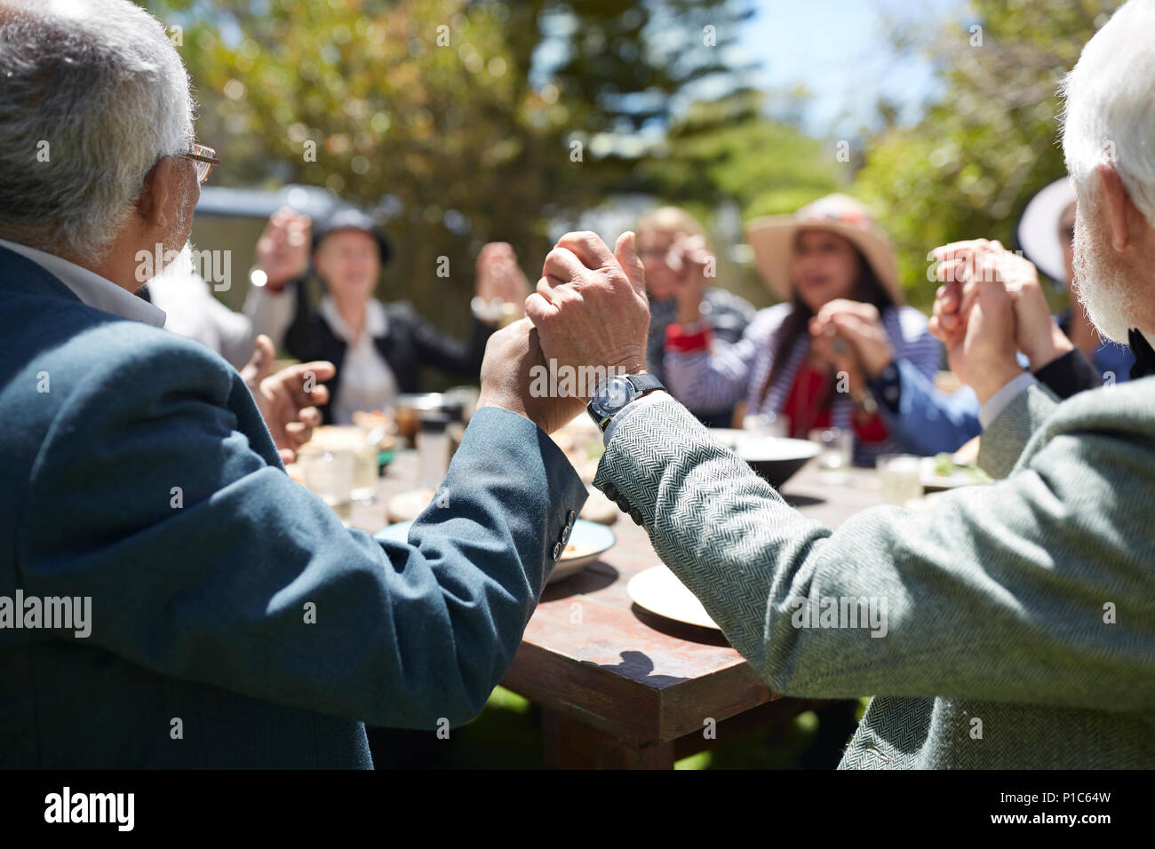 Active senior friends holding hands, praying at sunny garden party table Stock Photo