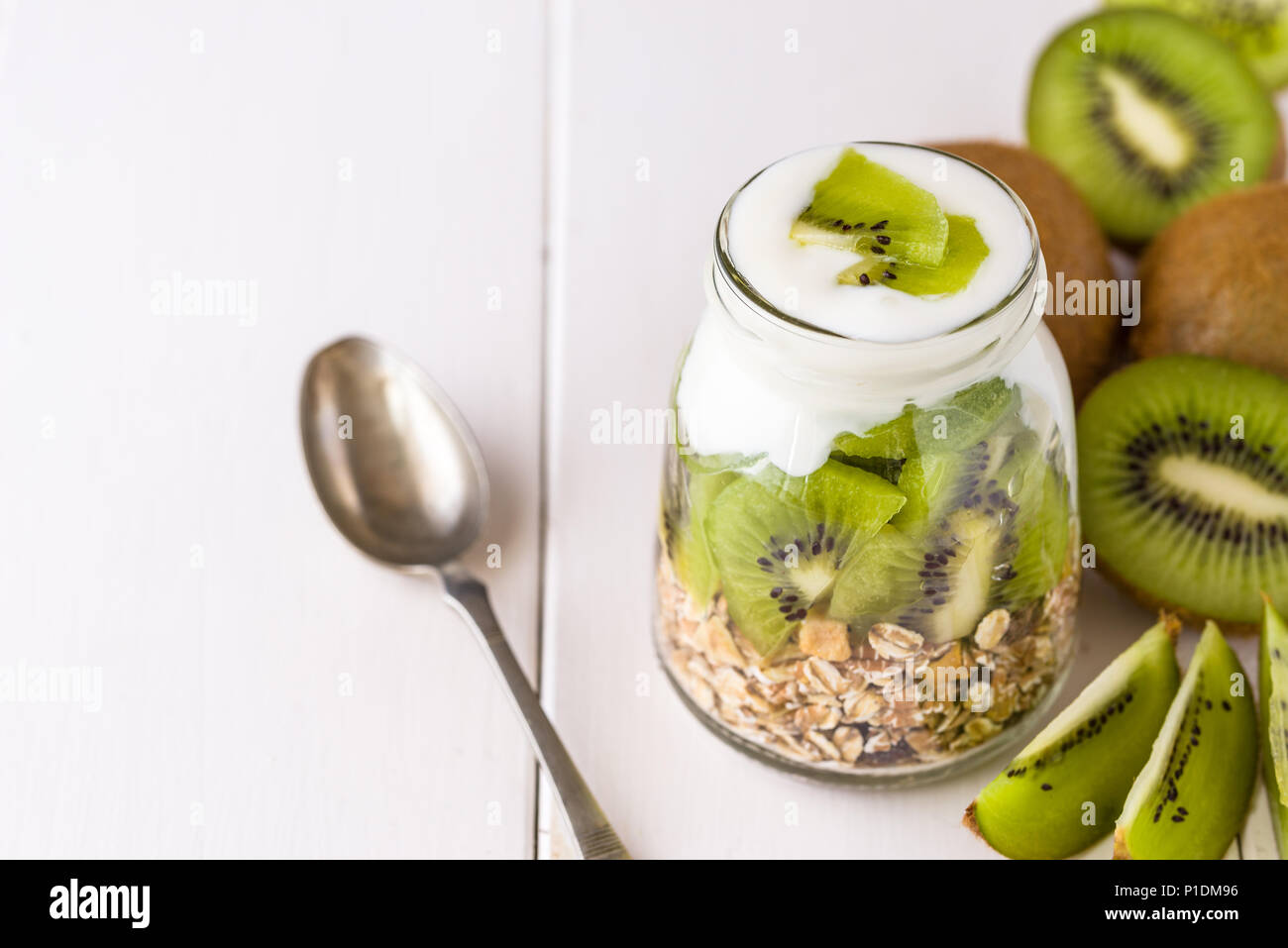 White yogurt with muesli in glass bowl with pieces of kiwi on top and whole pieces on the right side. Stock Photo