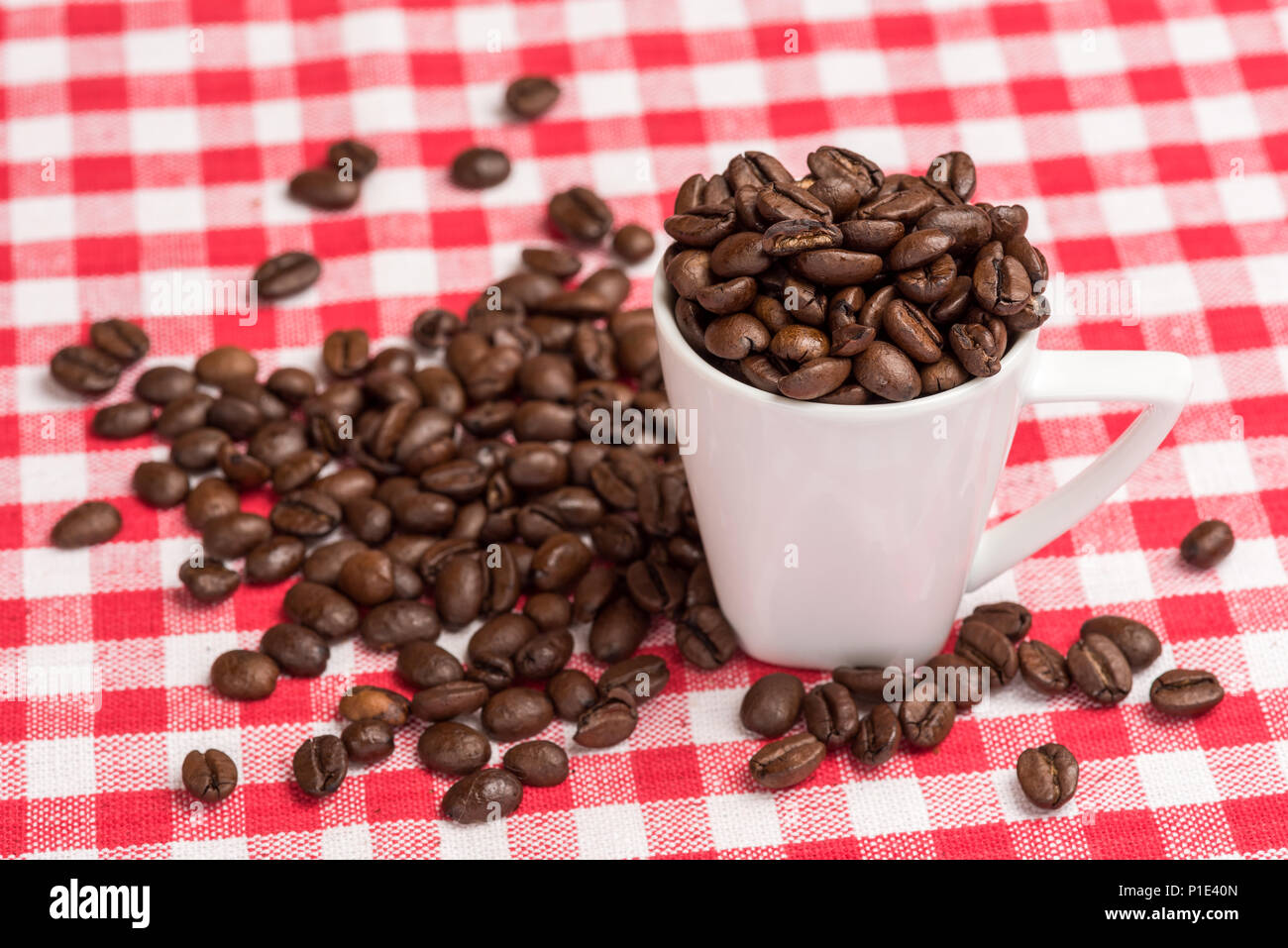 A white coffee mug is  filled with brown coffee beans. The coffee beans are also spread out on the kitchen table with a red checkered tablecloth as a  Stock Photo