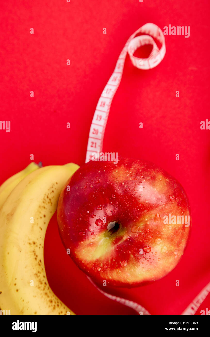 Measuring tape wrapped around a red apple and banana as a symbol of diet on red paper background. Weight loss concept. Diet. Dieting concept. Vegan. C Stock Photo