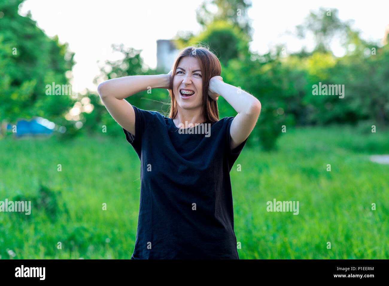 Girl emotionally covered her ears with her hands. Strong Scream unbearable noise. The girl is unhappy with her mouth open on teeth of the braces. Emotions of indignation. In summer park fresh air. . Stock Photo