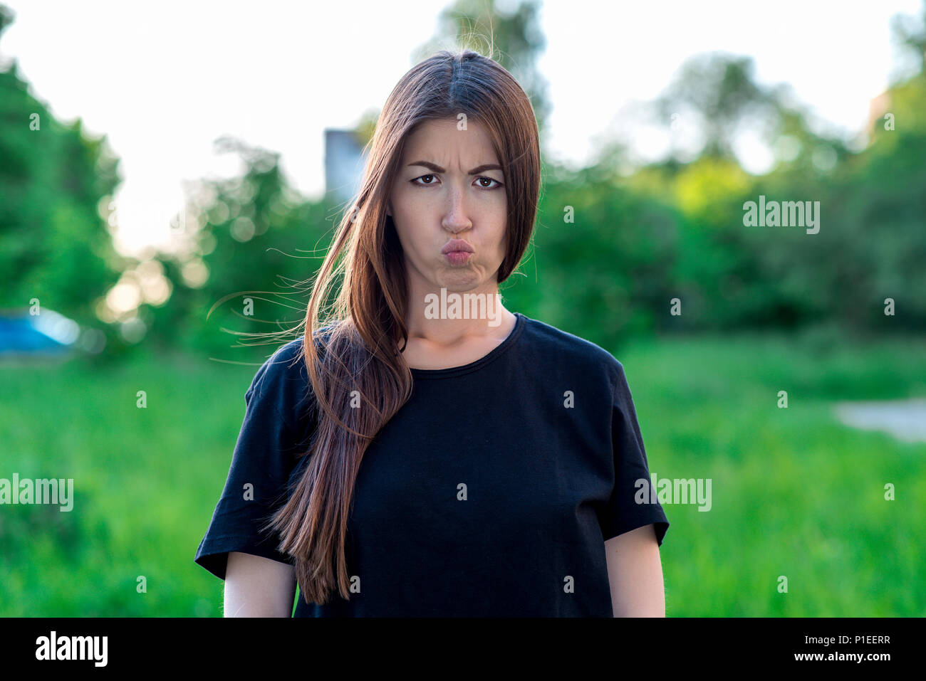 Girl in summer in the park in fresh air. Close-up portrait. The girl pouted. She puffed out her cheeks. Dissatisfied expression. Brunette with freckles. Emotional kiss. Whimsical Woman. Stock Photo
