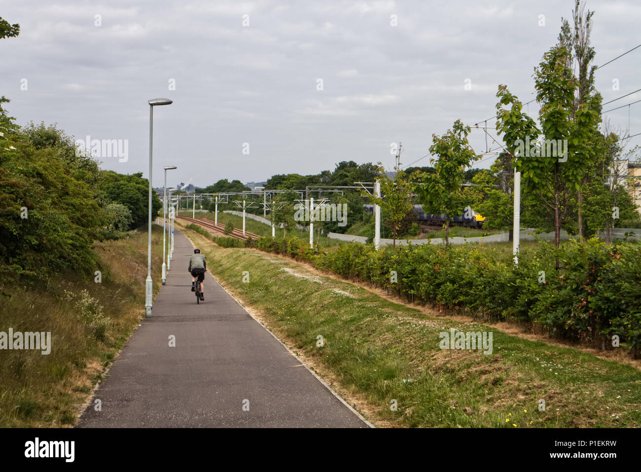Edinburgh, Scotland / United Kingdom - June 11 2018: A man is cycling down a slope alongside a tram track while a Scotland train is travelling in the Stock Photo