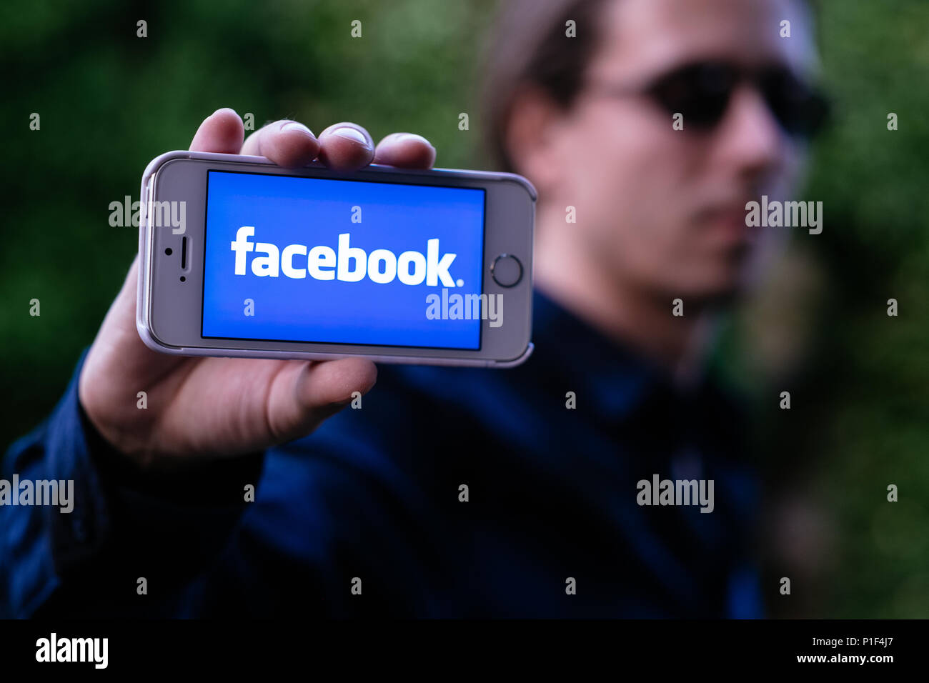 Closeup of serious young man with sunglasses holding white iPhone with FACEBOOK LOGO on screen Stock Photo