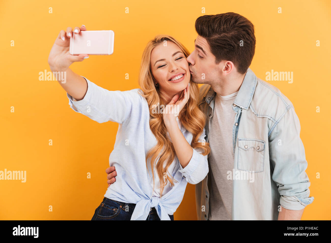 Portrait of lovely couple taking selfie photo on mobile phone while man kissing woman on cheek isolated over yellow background Stock Photo