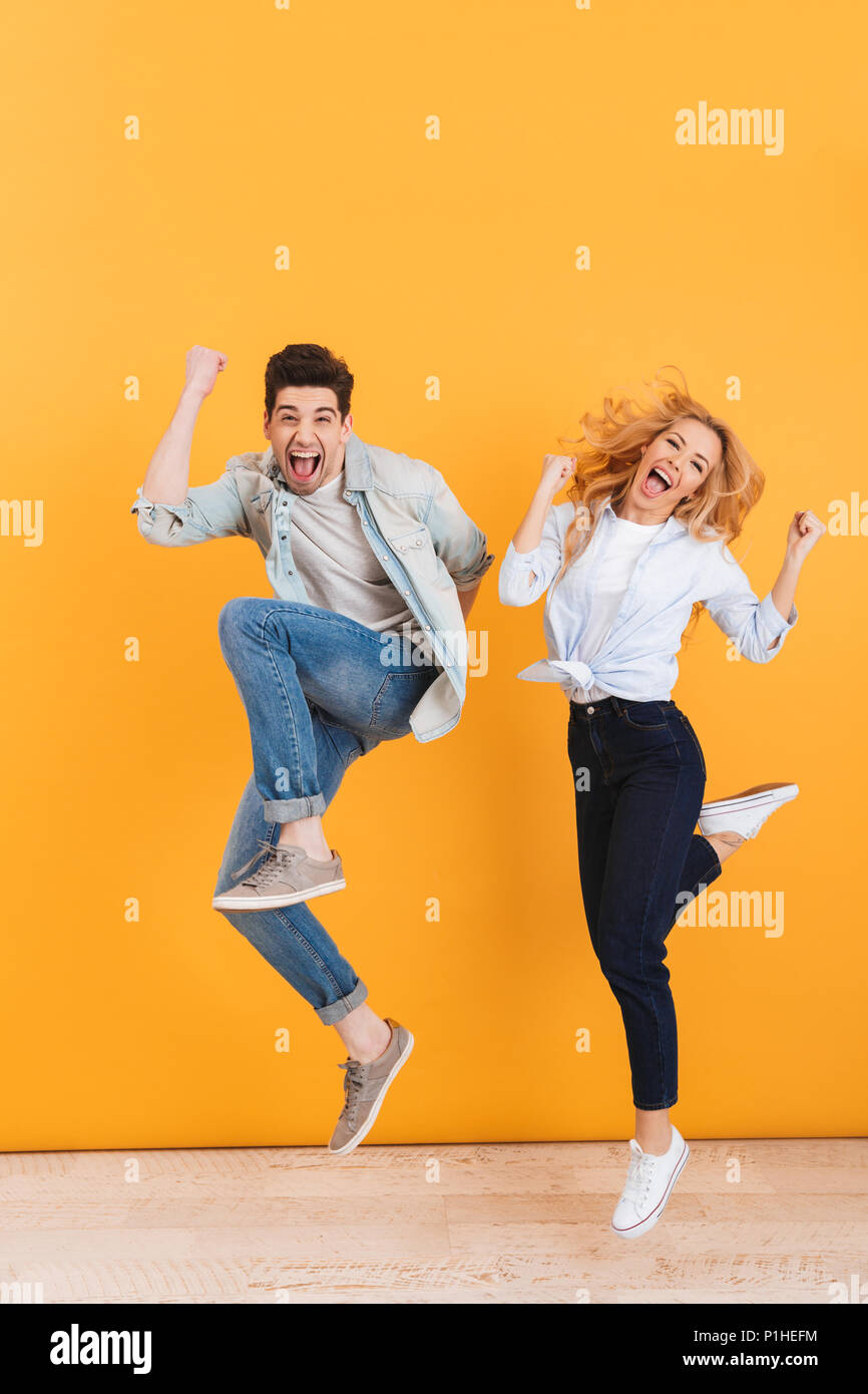 Full length photo of happy young couple screaming in surprise while jumping and clenching fists like winners isolated over yellow background Stock Photo