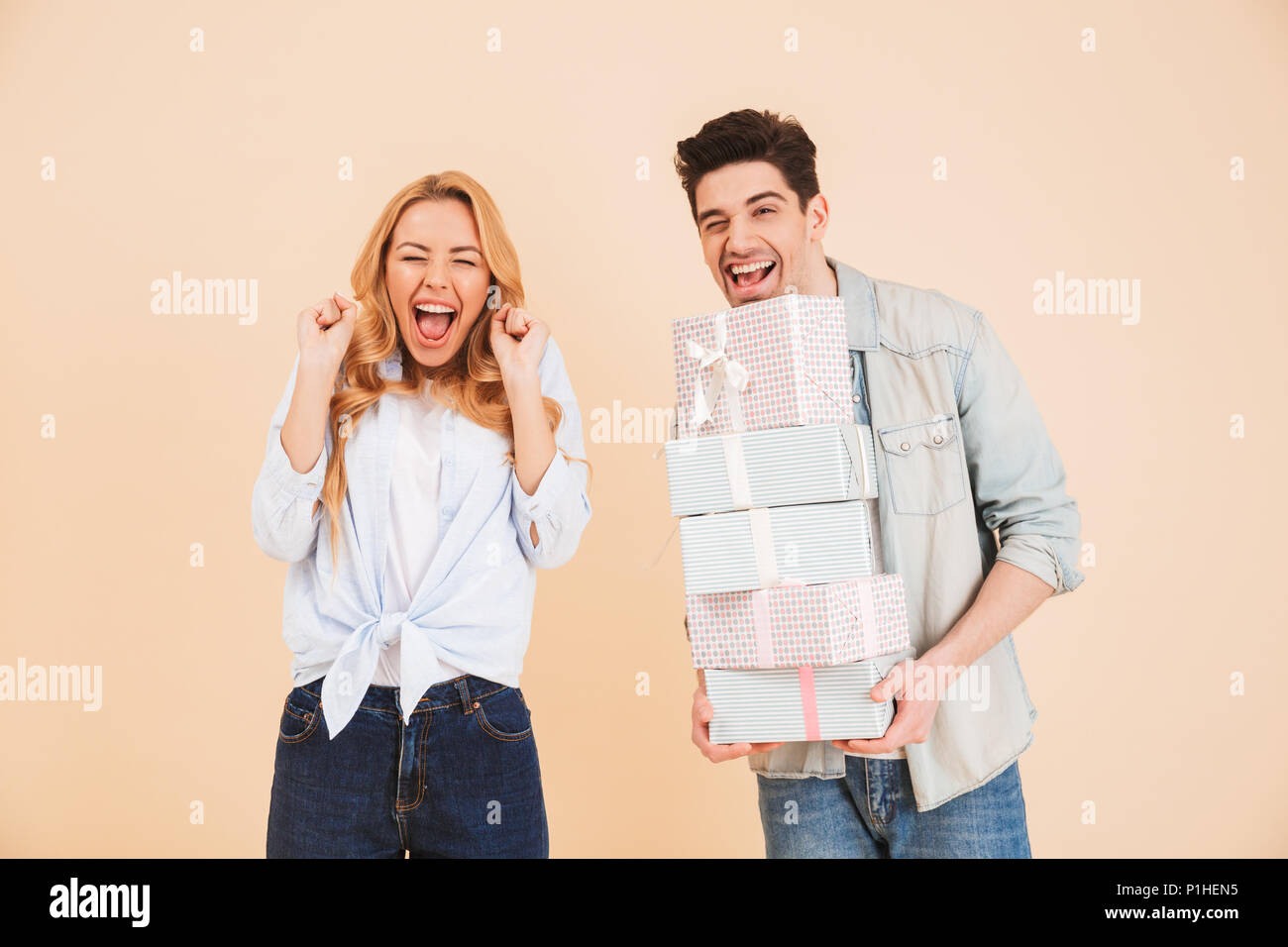 Image of surprised woman shouting in happiness while handsome man holding lots of gift boxes isolated over beige background Stock Photo