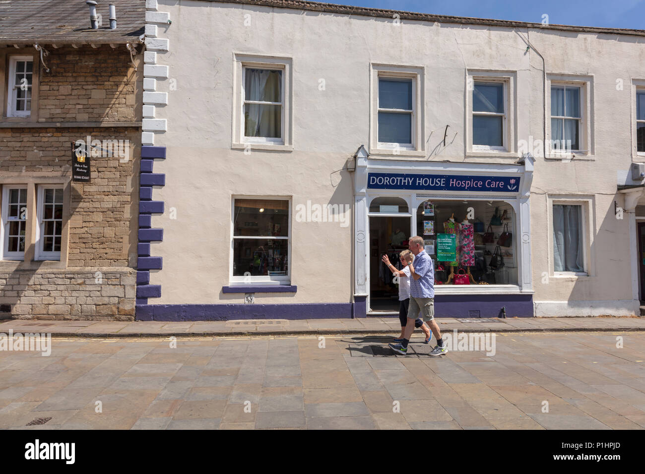 A couple walk past The Dorothy House Hospice charity shop on the Market Square, Malmesbury, Wiltshire, UK. The man is vaping. Stock Photo