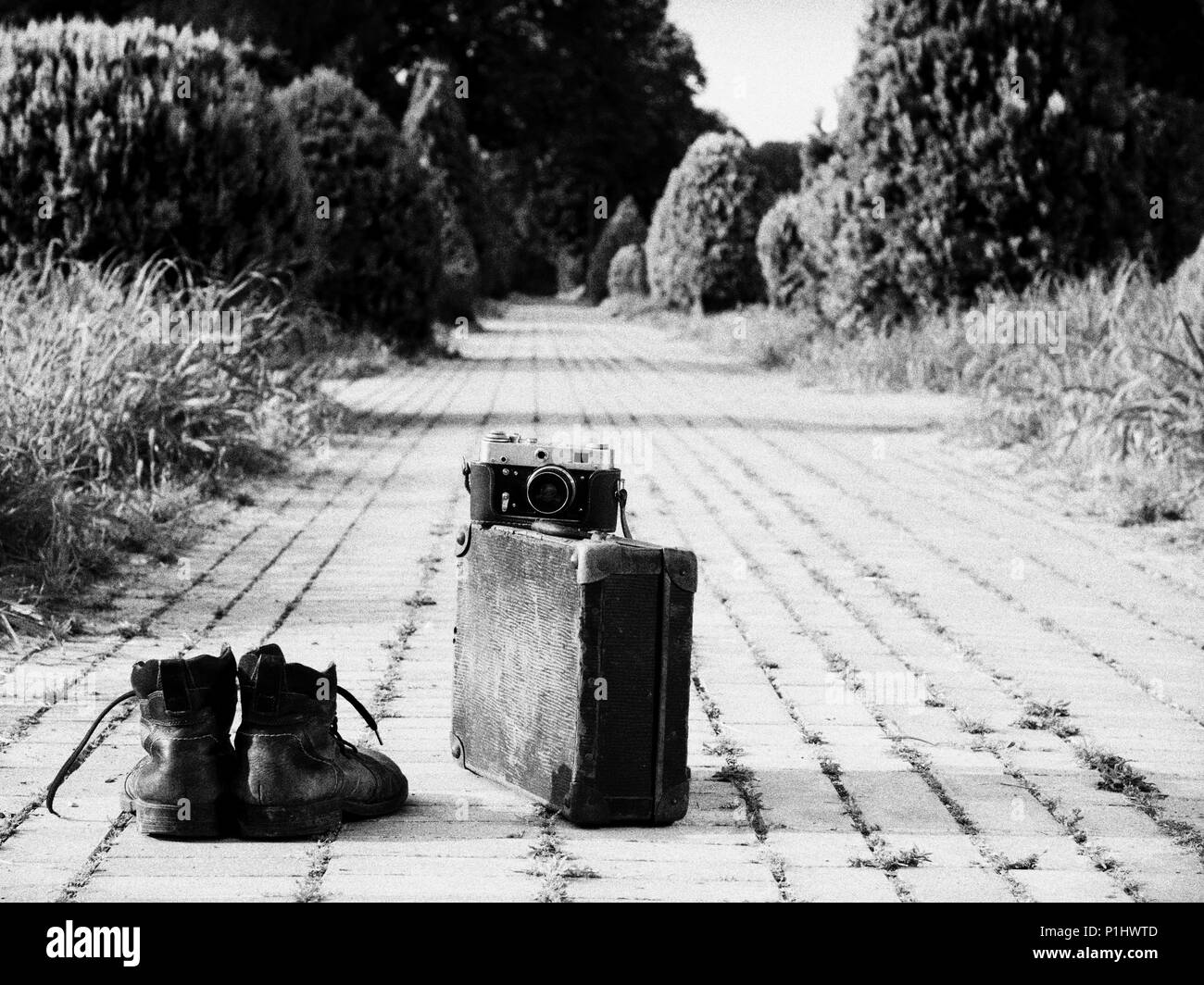 Leather boots, a vintage suitcase, and a film camera in its open leather case, in the middle of a brick road. A B/W photo. Grain noise added. Stock Photo