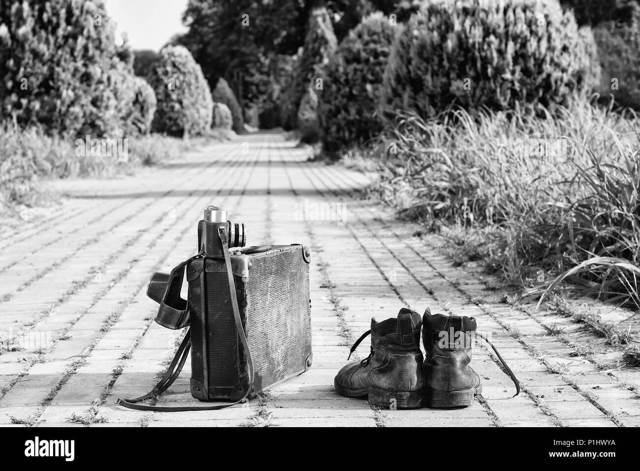 Traveling light! Worn ankle boots next to a vintage cardboard suitcase, a film camera in its open leather case, and a brick road. A B/W photo effect. Stock Photo