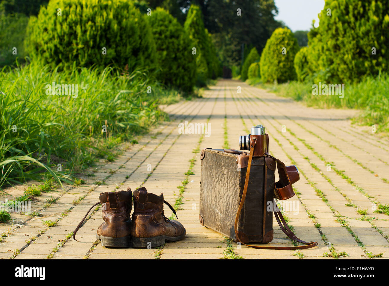 Traveling light! Shabby ankle boots next to a vintage cardboard suitcase, a film camera in its open leather case, and a yellow brick road. Stock Photo