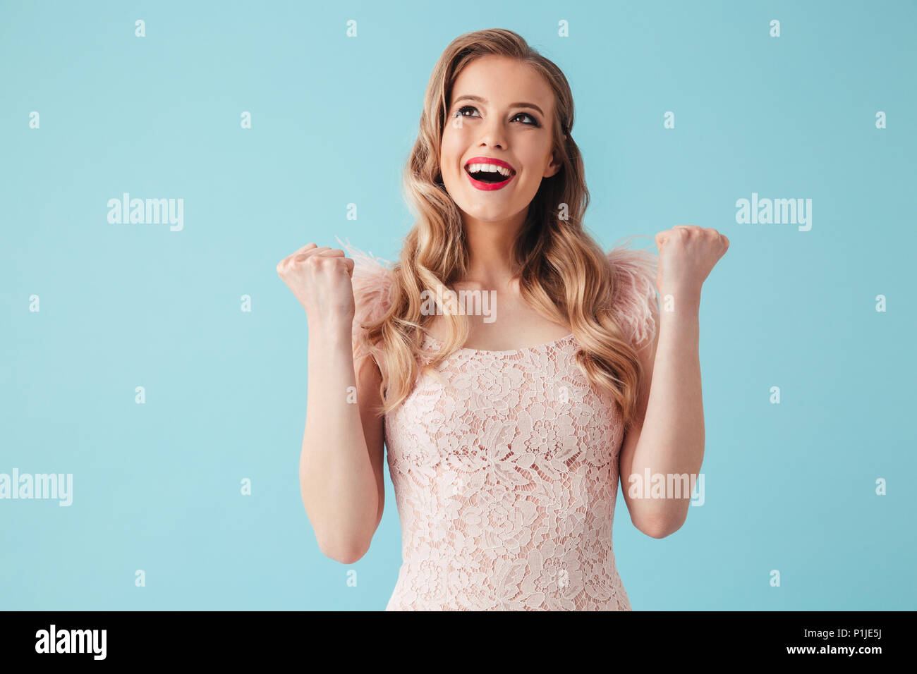 Cheerful blonde woman in dress rejoices and looking up over turquoise background Stock Photo