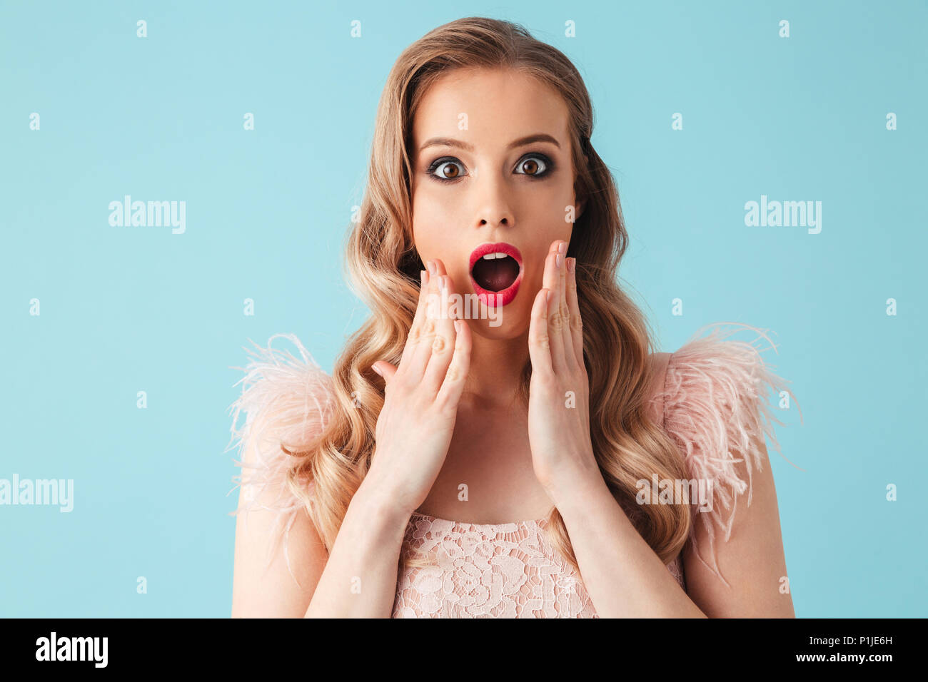 Surprised blonde woman in dress touching her cheeks and looking at the camera over turquoise background Stock Photo