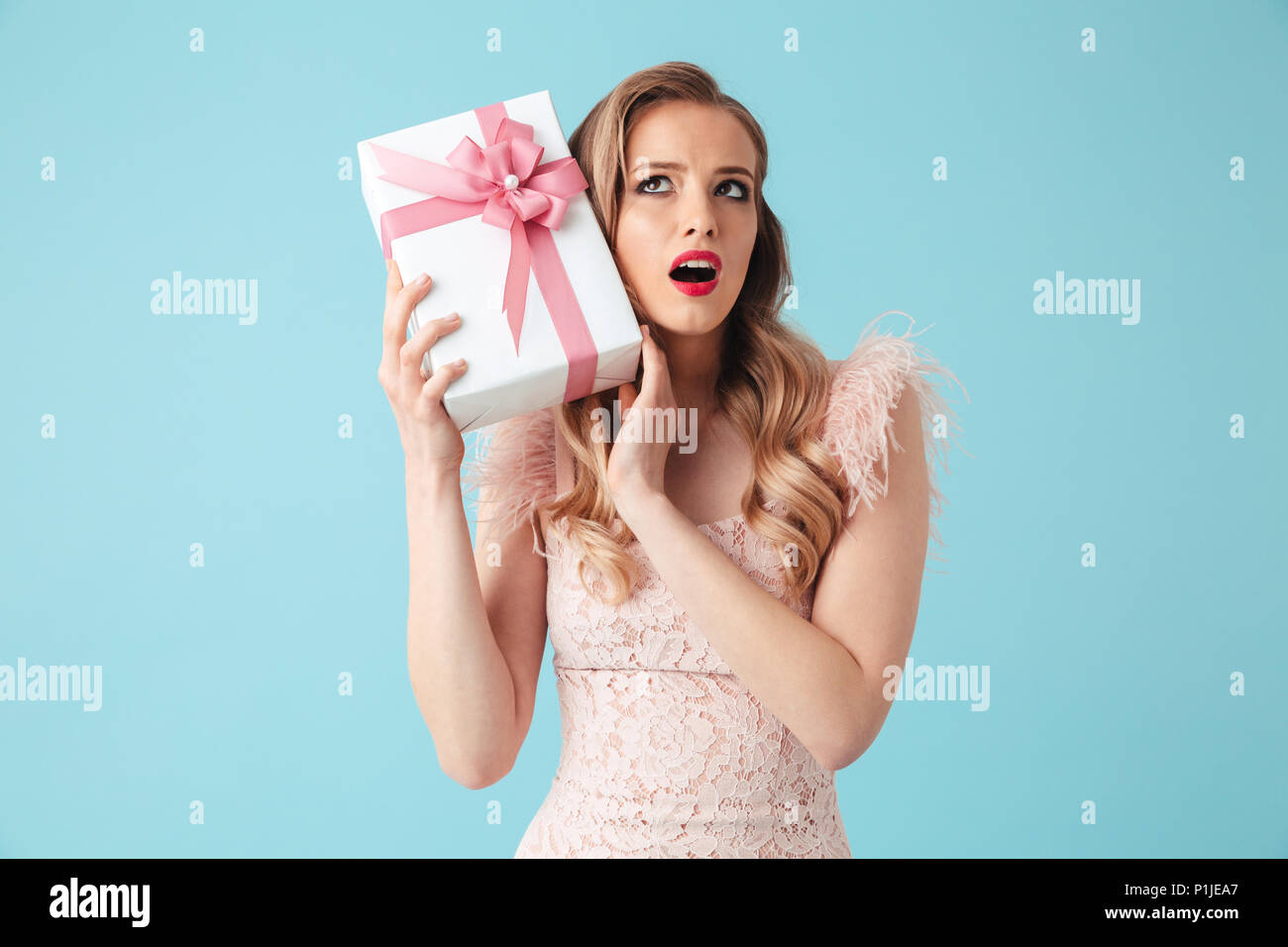 Intrigued blonde woman in dress holding gift box and looking up over turquoise background Stock Photo