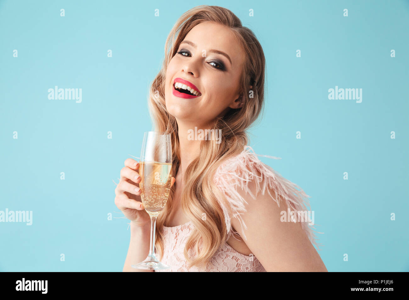 Side view of Cheerful blonde woman in dress posing with champagne and looking at the camera over turquoise background Stock Photo