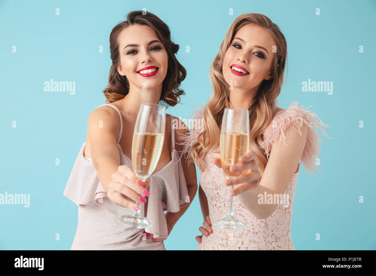 Two happy women in dresses posing together drinking champagne and looking at the camera over turquoise background Stock Photo