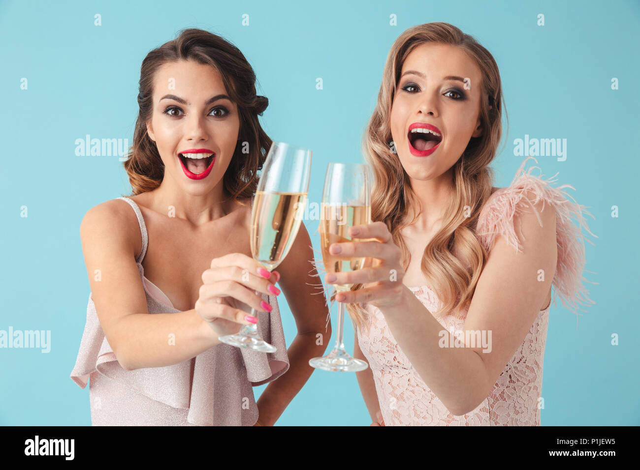 Two surprised happy women in dresses drinking champagne and looking at the camera over turquoise background Stock Photo