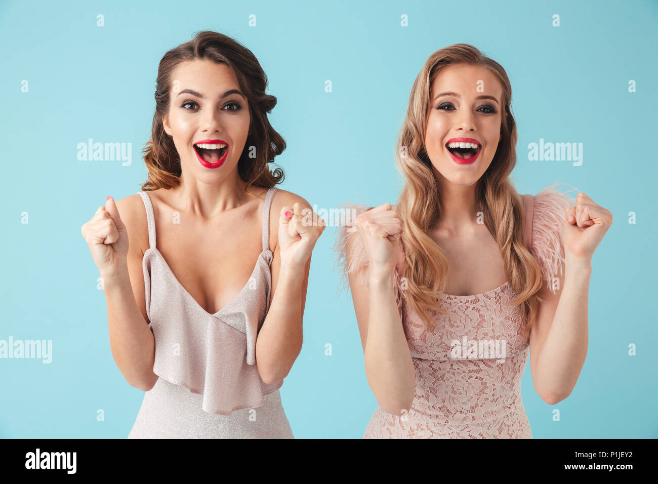 Two Surprised happy women in dresses screaming and rejoices while looking at the camera over turquoise background Stock Photo