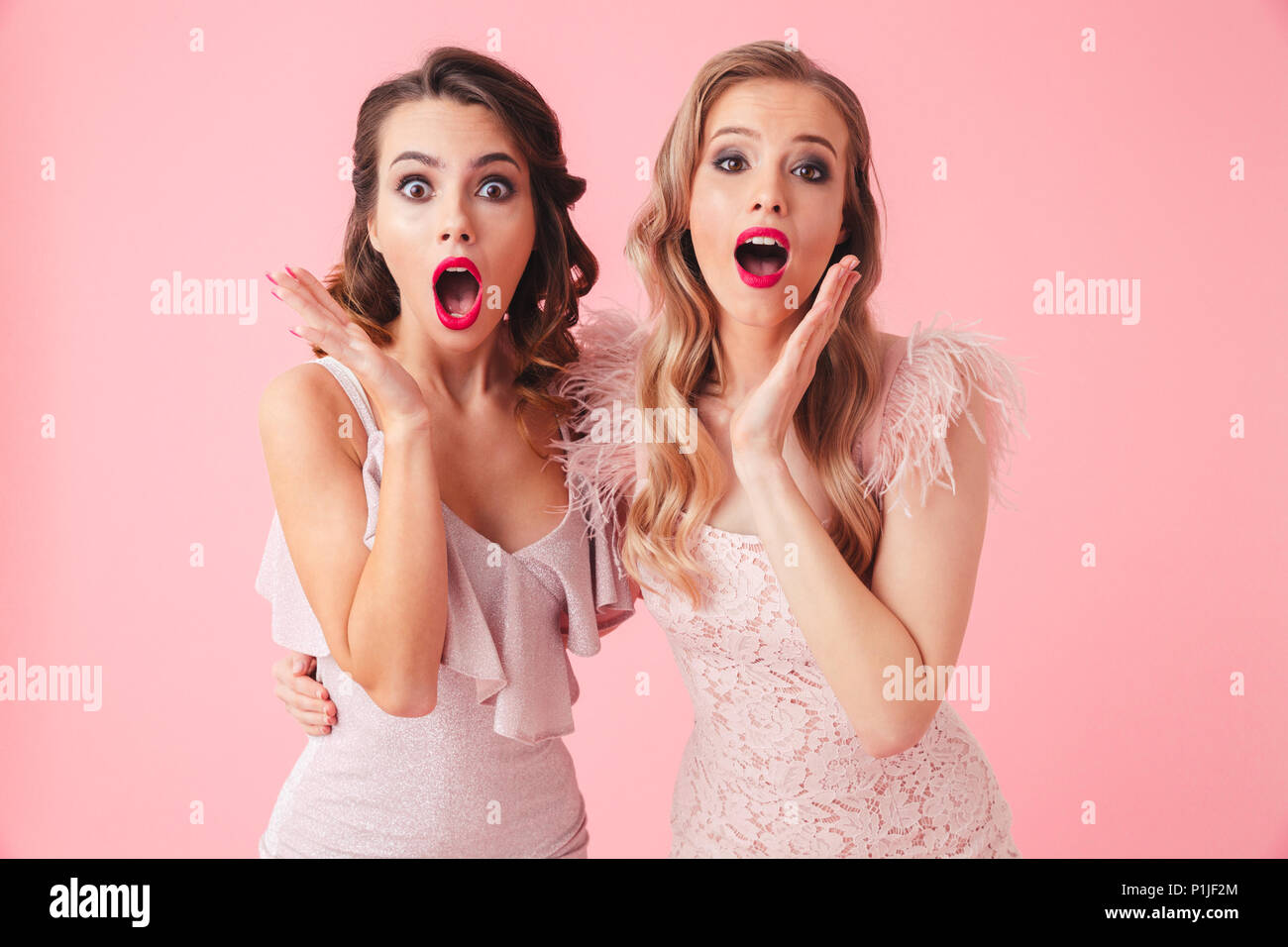 Two shocked women in dresses hugging to each other and looking at the camera with open mouth over pink background Stock Photo