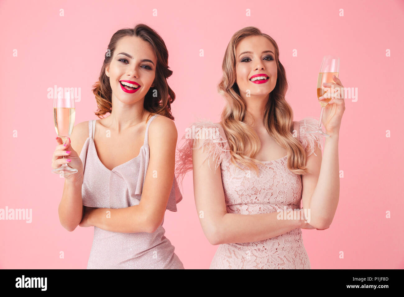 Two Happy elegant women in dresses posing together with champagne and looking at the camera over pink background Stock Photo
