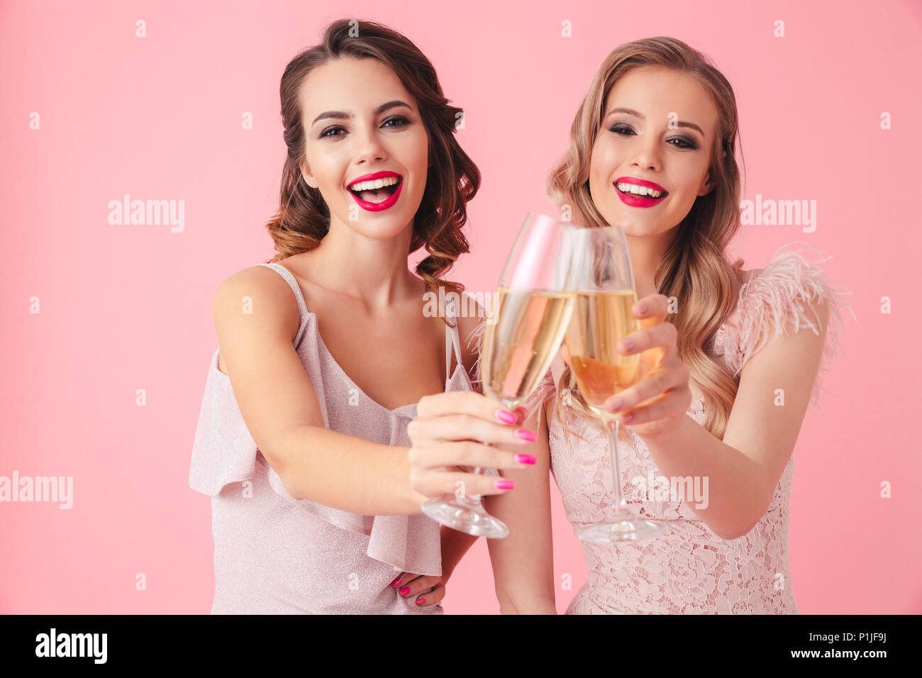 Two cheerful women in dresses drinking champagne and looking at the camera over pink background Stock Photo