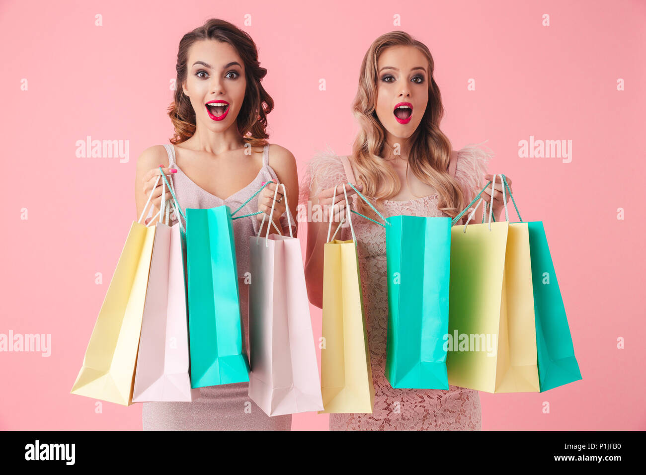 Two surprised happy women in dresses opening packages and looking at the camera over pink background Stock Photo