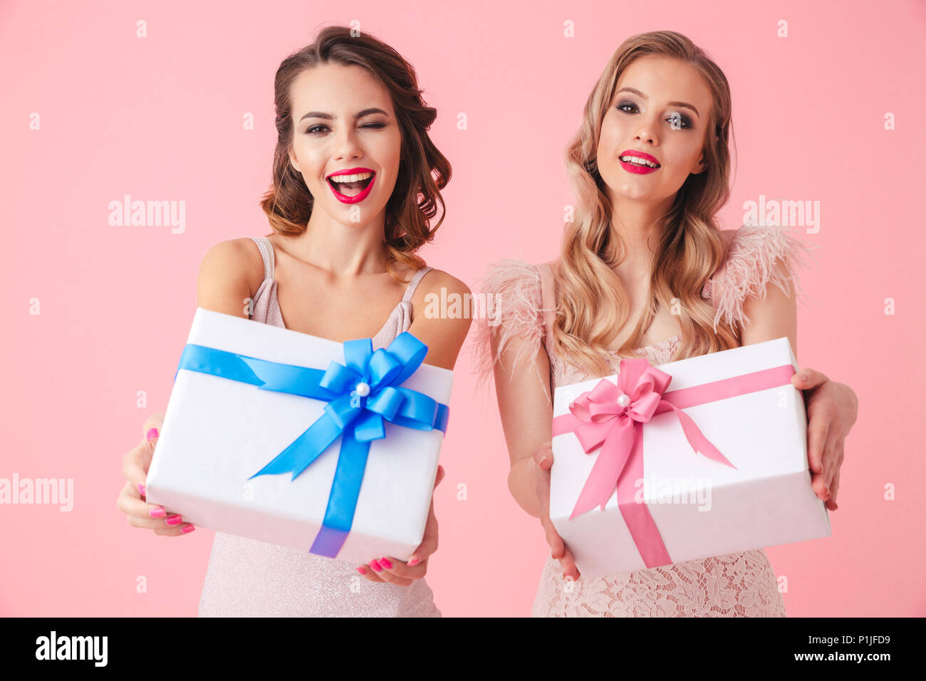 Two cheerful women in dresses having fun together while holding big gift boxes and looking at the camera over pink background Stock Photo